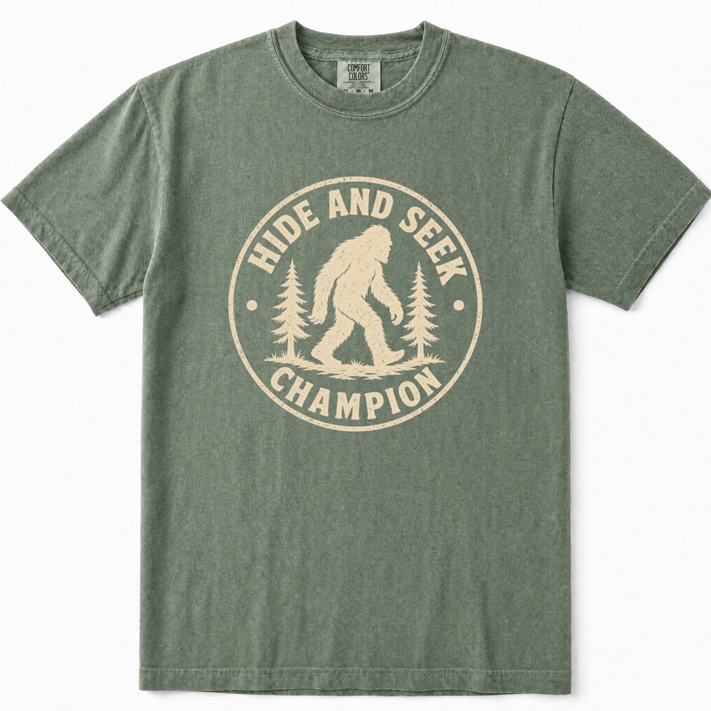 Moss green crewneck t-shirt featuring a distressed beige circular graphic on the front with the text 'HIDE AND SEEK CHAMPION' and an illustration of a Bigfoot figure between pine trees.