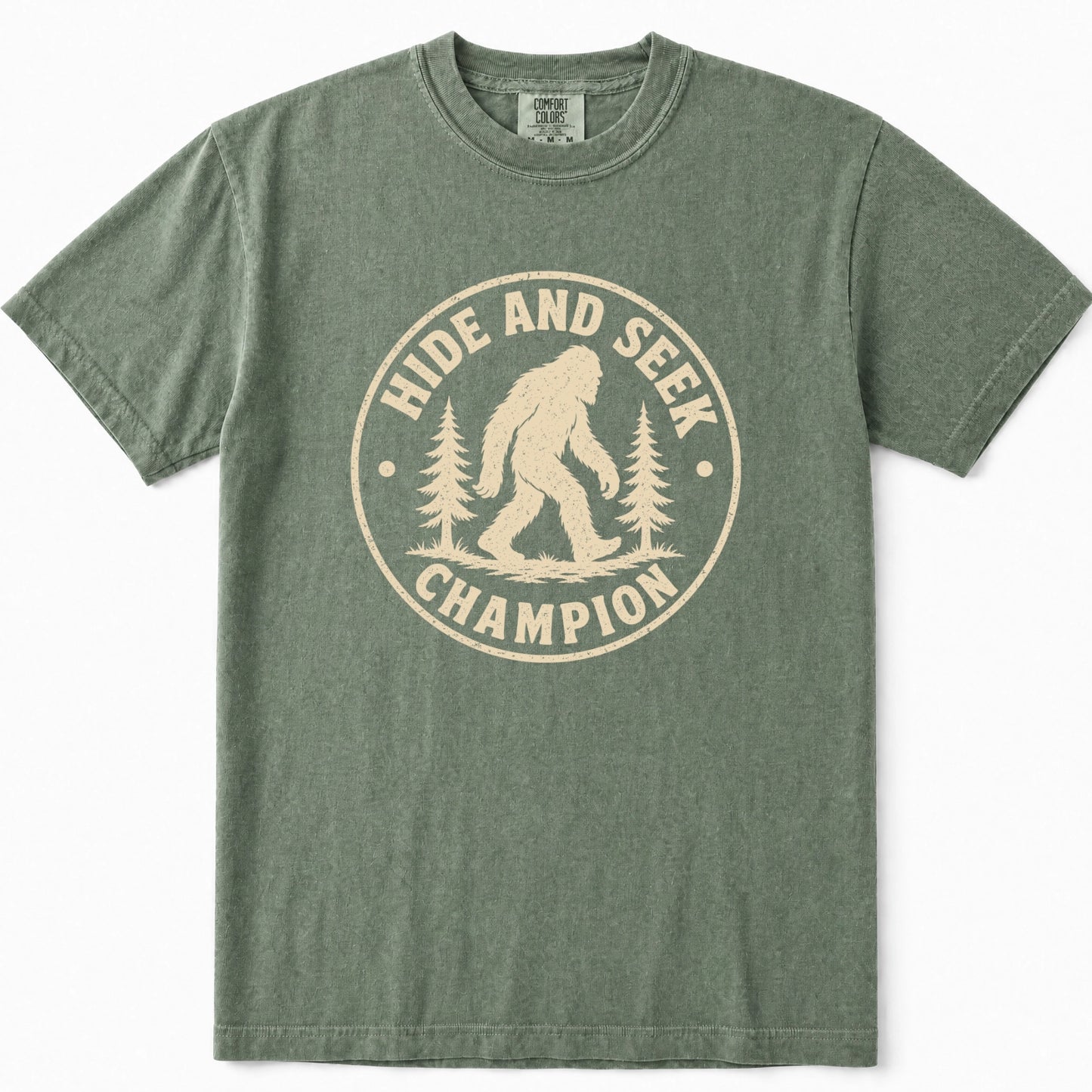 Moss green crewneck t-shirt featuring a distressed beige circular graphic on the front with the text 'HIDE AND SEEK CHAMPION' and an illustration of a Bigfoot figure between pine trees.