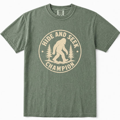 Moss green crewneck t-shirt featuring a distressed beige circular graphic on the front with the text 'HIDE AND SEEK CHAMPION' and an illustration of a Bigfoot figure between pine trees.