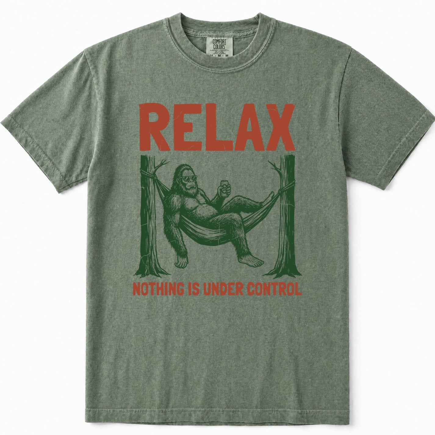 Moss green t-shirt featuring a graphic of Bigfoot lounging in a hammock between two trees, with the word 'RELAX' printed in orange-red above and 'NOTHING IS UNDER CONTROL' below.
