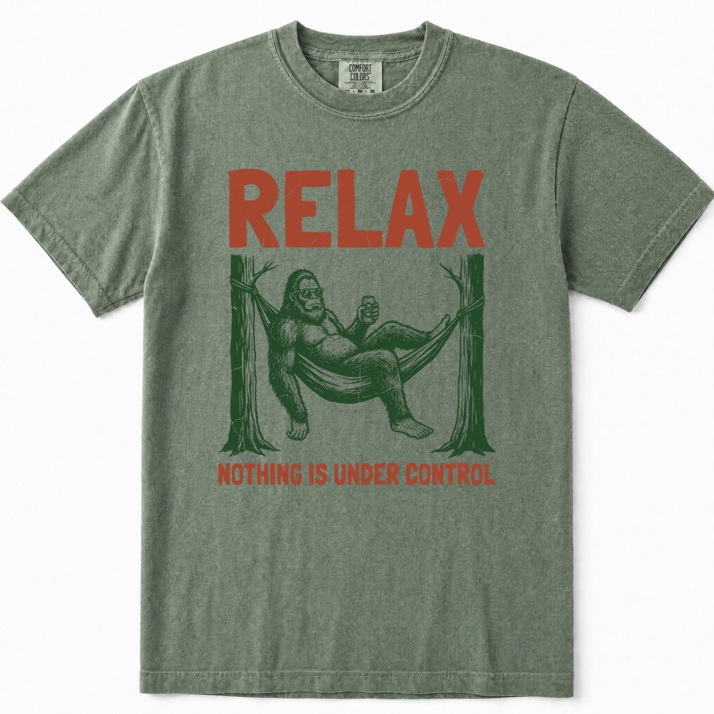 Moss green t-shirt featuring a graphic of Bigfoot lounging in a hammock between two trees, with the word 'RELAX' printed in orange-red above and 'NOTHING IS UNDER CONTROL' below.