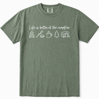 Moss green short-sleeve t-shirt with a white graphic print on the chest that reads 'Life is better at the campfire' above simple camping icons including a campfire, marshmallow, mug, tree, and camper. The shirt is displayed flat against a clean white background.