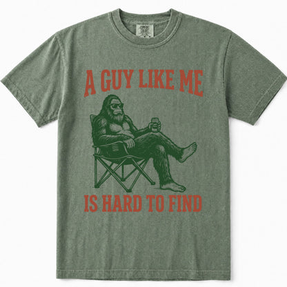 Moss green t-shirt featuring a graphic of Bigfoot sitting in a folding chair holding a beer, with the text 'A GUY LIKE ME' and 'IS HARD TO FIND' printed in a reddish-orange color.