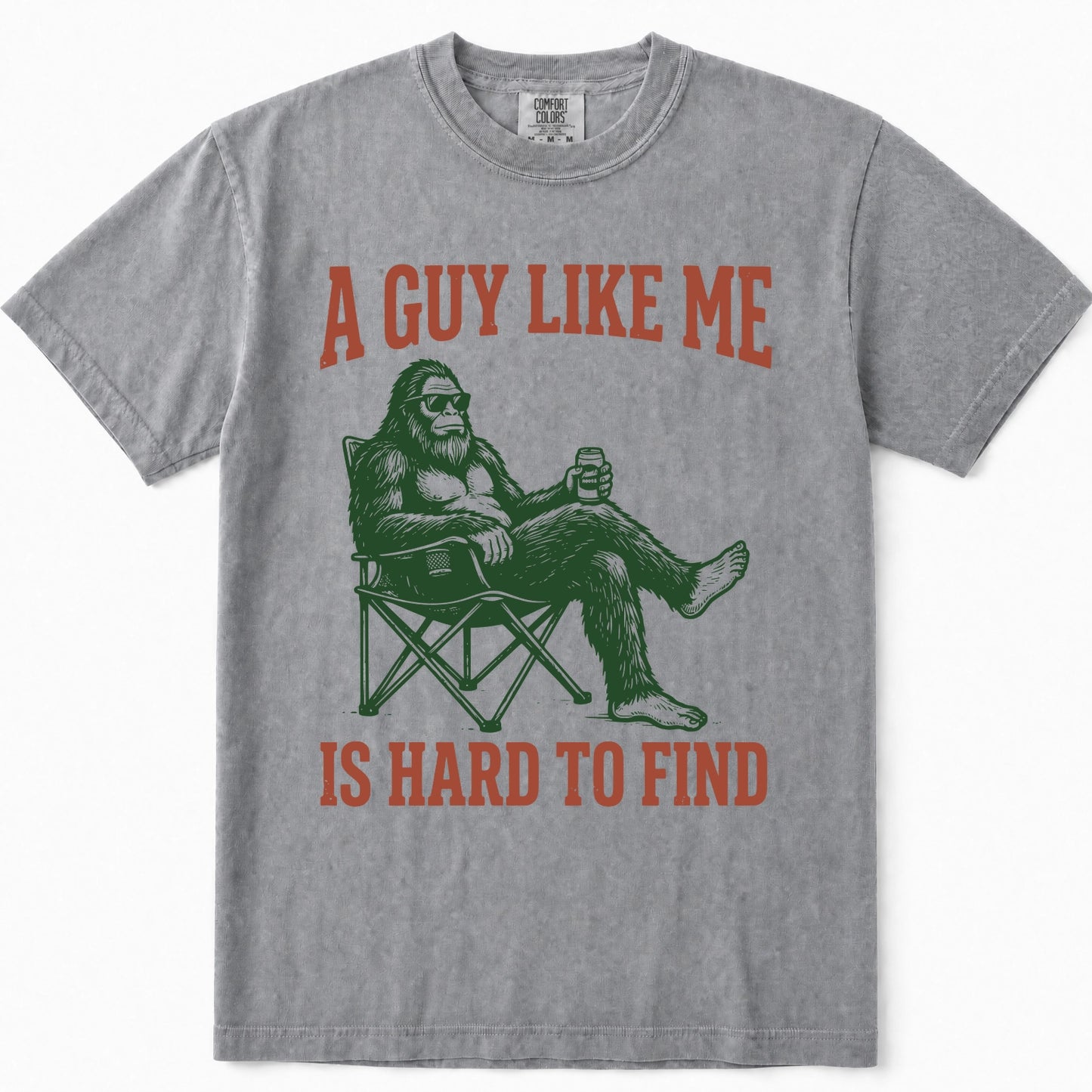 Grey short-sleeve t-shirt with a graphic print of Bigfoot sitting in a folding chair holding a beer, with the text 'A GUY LIKE ME IS HARD TO FIND' in distressed orange-red lettering.