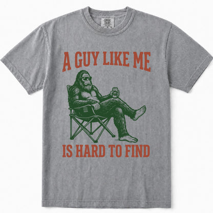 Grey short-sleeve t-shirt with a graphic print of Bigfoot sitting in a folding chair holding a beer, with the text 'A GUY LIKE ME IS HARD TO FIND' in distressed orange-red lettering.