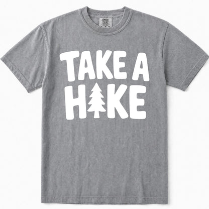 Grey short-sleeve crewneck t-shirt with a bold white graphic print on the front that reads 'TAKE A HAKE' with a pine tree icon replacing the letter 'I'.