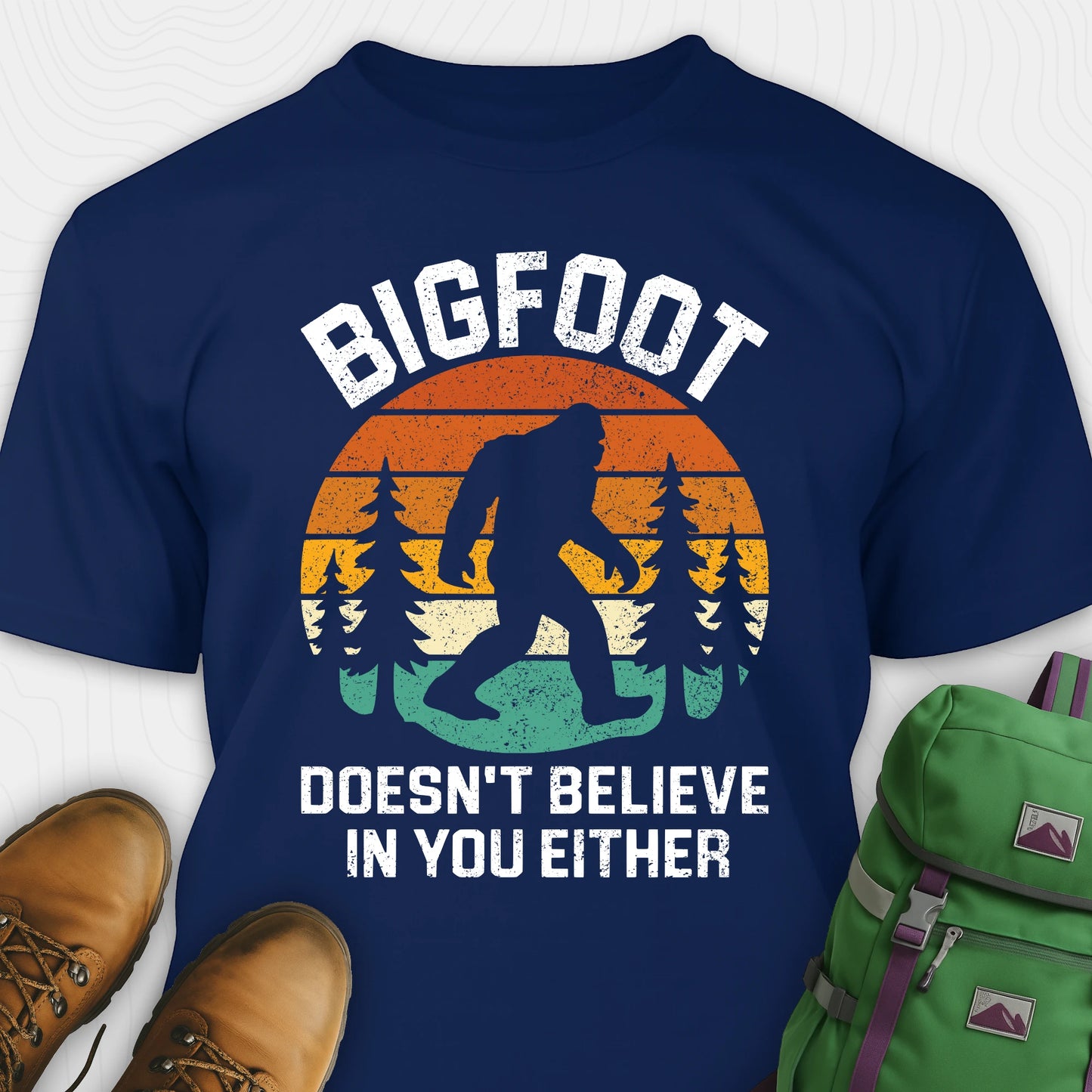 Navy funny Bigfoot shirt with sasquatch silhouette and humorous saying, paired with brown boots and a green backpack on a white background.
