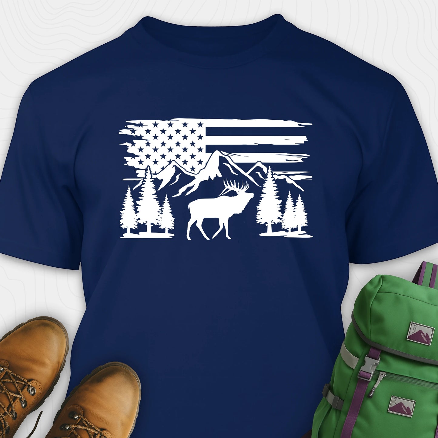 Navy deer t-shirt with black graphic design of an American flag, trees, and a moose, accompanied by brown boots and a green backpack.