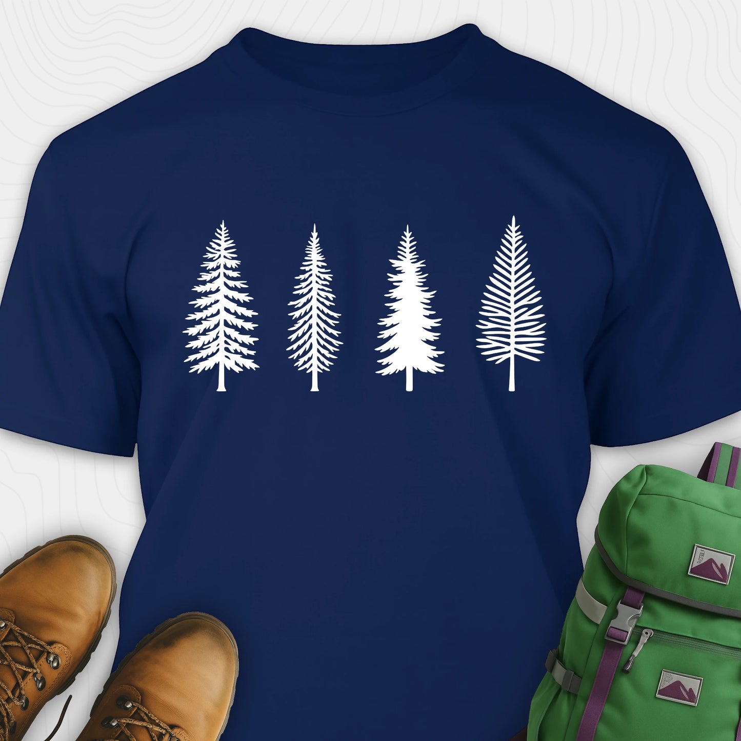 Navy pine tree shirt with white minimalist forest graphic, brown boots, and a green backpack on a light gray background