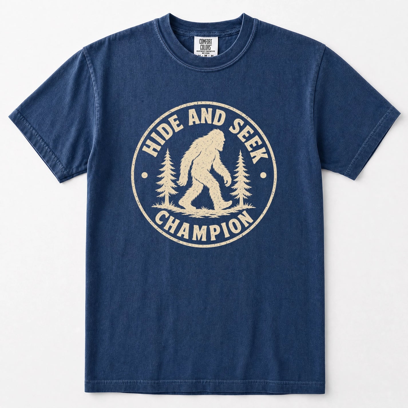 Navy blue short-sleeve t-shirt with a distressed cream-colored circular graphic on the chest that reads 'HIDE AND SEEK CHAMPION' with a Bigfoot silhouette and pine trees.