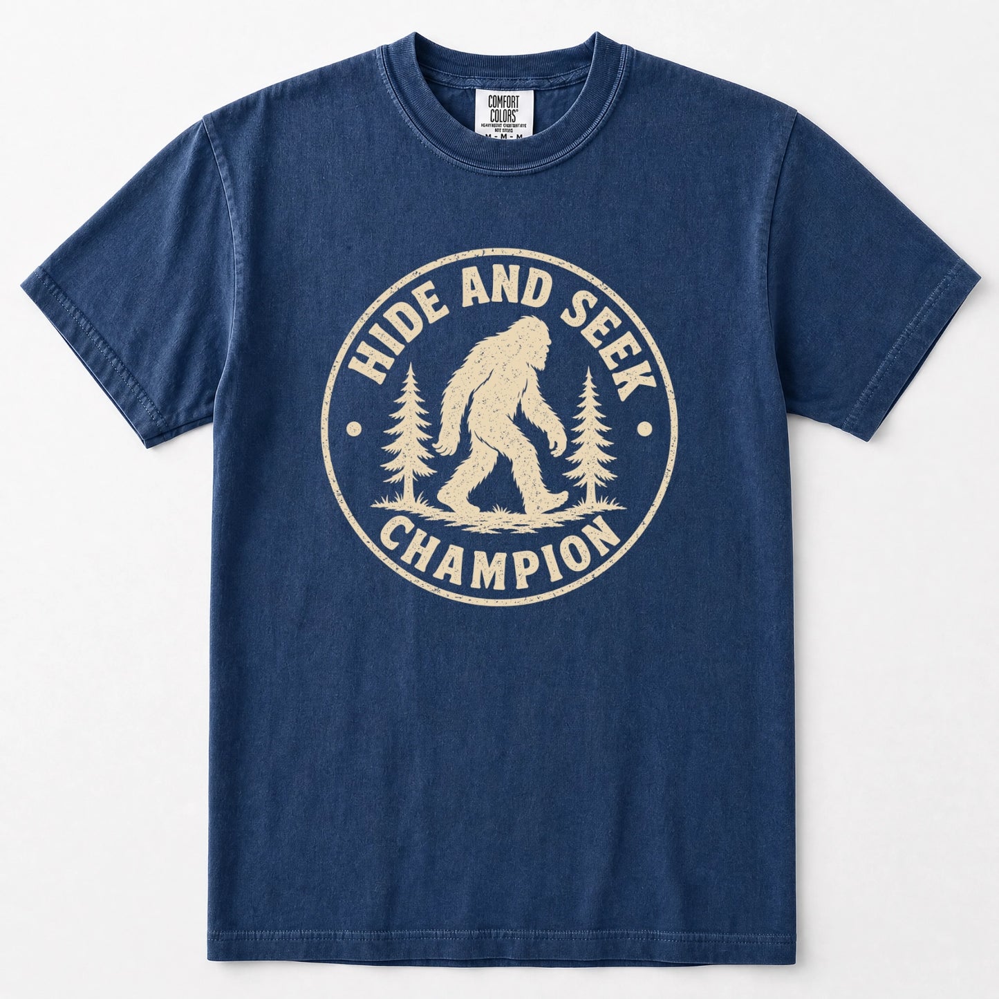 Navy blue short-sleeve t-shirt with a distressed cream-colored circular graphic on the chest that reads 'HIDE AND SEEK CHAMPION' with a Bigfoot silhouette and pine trees.