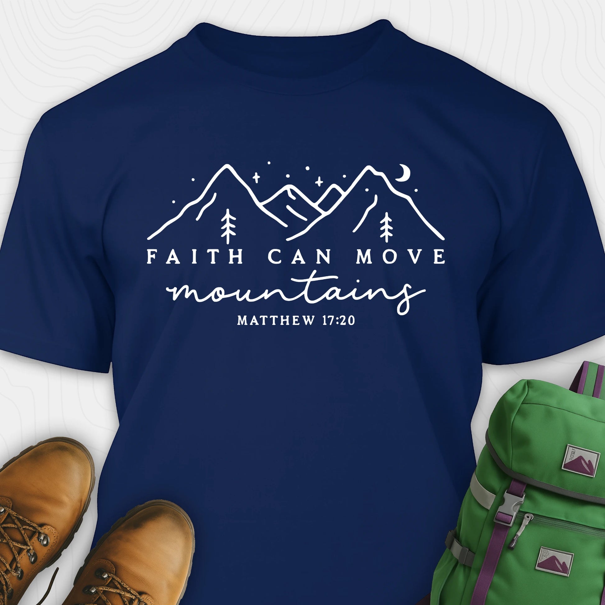 Navy faith can move mountains shirt with mountain scripture design, paired with hiking boots and a green backpack.