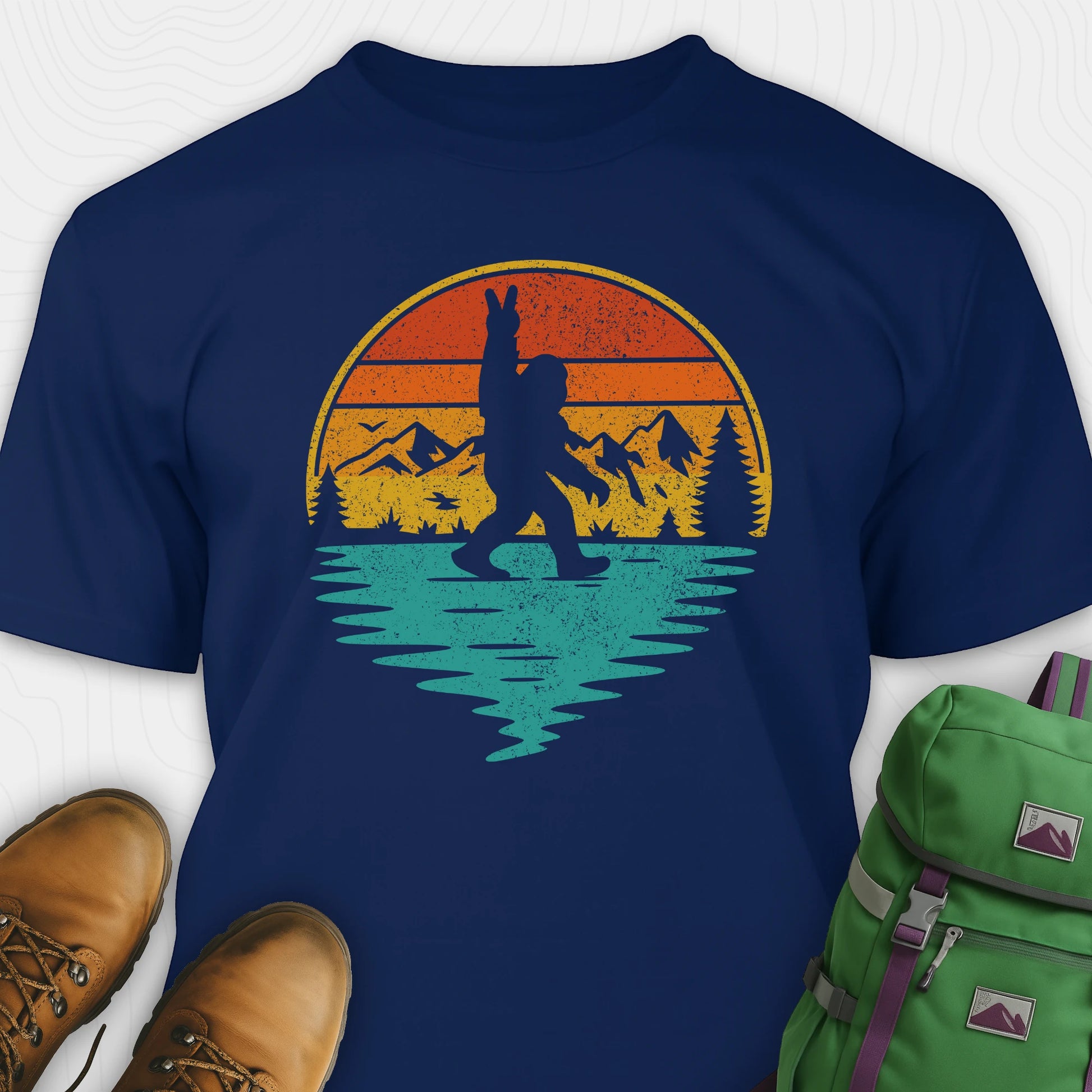 Navy bigfoot shirt for men with retro mountain graphic beside boots and green backpack