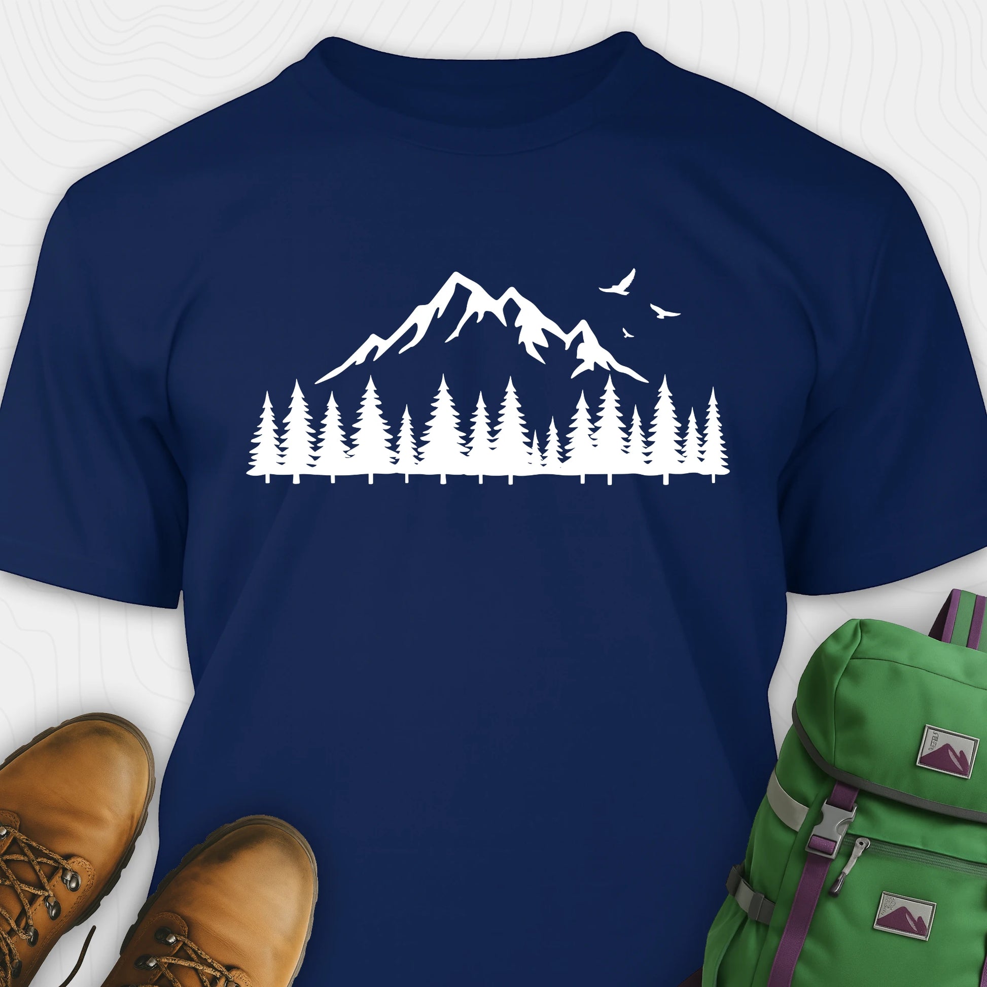 Navy wild t-shirt with a minimalist mountain and pine tree graphic, shown flat with boots and a backpack