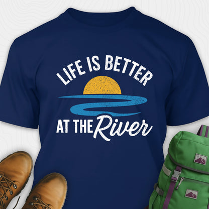 Navy river shirt with 'Life is Better at the River' design, paired with brown boots and a green backpack on a light gray background.