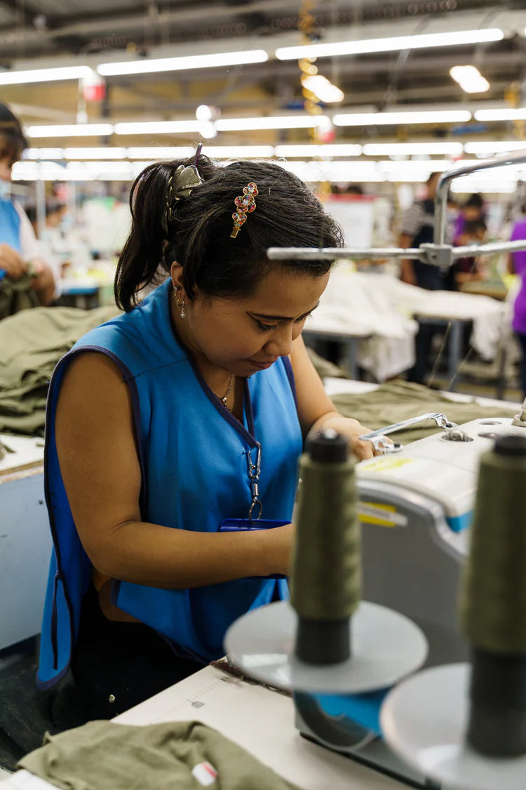 A woman wearing a blue vest sits at an industrial sewing machine in a garment factory, looking down with focus as she stitches olive-green fabric. Large spools of green thread are prominent in the foreground, while the background shows a brightly lit, busy workshop with rows of lights.