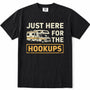 Black short-sleeve crewneck t-shirt with a vintage-style graphic print on the front. The graphic features the text 'JUST HERE FOR THE HOOKUPS' in distressed cream and yellow lettering, accompanied by an illustration of a cream and yellow camper van.