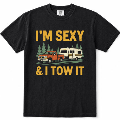 Black short-sleeve crewneck t-shirt with a vintage-style graphic print that reads 'I'M SEXY & I TOW IT' in yellow text, featuring an illustration of an orange pickup truck towing a cream camper trailer with green pine trees.