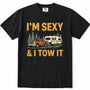 Black short-sleeve crewneck t-shirt with a vintage-style graphic print that reads 'I'M SEXY & I TOW IT' in yellow text, featuring an illustration of an orange pickup truck towing a cream camper trailer with green pine trees.