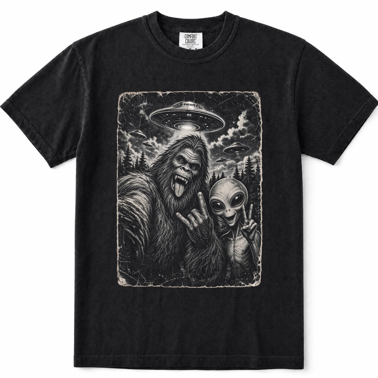 Black short-sleeve crewneck t-shirt featuring a large rectangular graphic print on the front. The graphic is a monochrome illustration depicting a Bigfoot taking a selfie with an alien under a flying saucer.