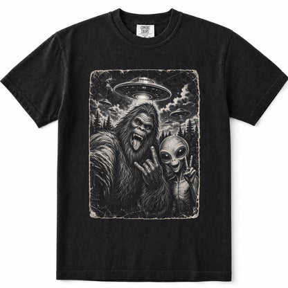 Black short-sleeve crewneck t-shirt featuring a large rectangular graphic print on the front. The graphic is a monochrome illustration depicting a Bigfoot taking a selfie with an alien under a flying saucer.