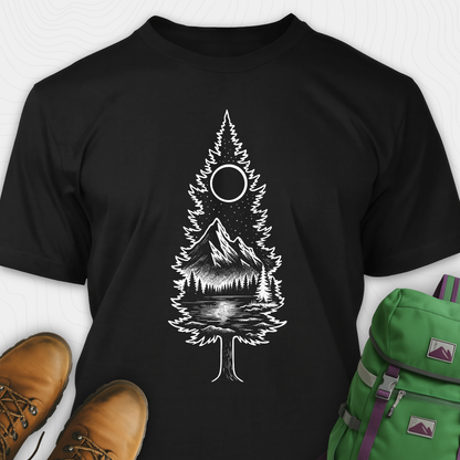 A black tree t-shirt featuring a detailed white graphic of a pine tree silhouette. The tree outline contains a landscape scene: a mountain peak rising over a forested shoreline, with its reflection visible in a lake below. Above the mountain is a starry night sky and a full or partial circle representing the moon or sun. The t-shirt is displayed flat with brown leather boots to the lower left and a green and purple backpack to the lower right, suggesting an outdoor or hiking theme.
