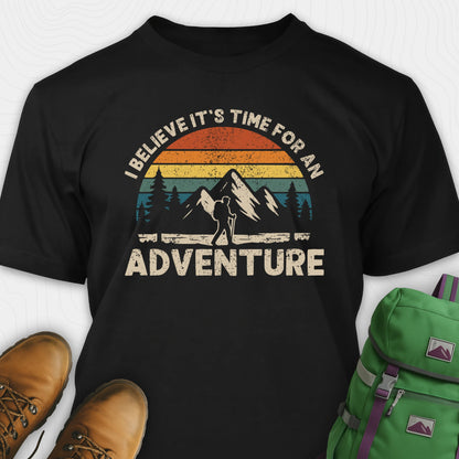 Black backpacking shirt with mountain graphic and 'I believe it's time for an adventure' text, paired with brown boots and a green backpack.