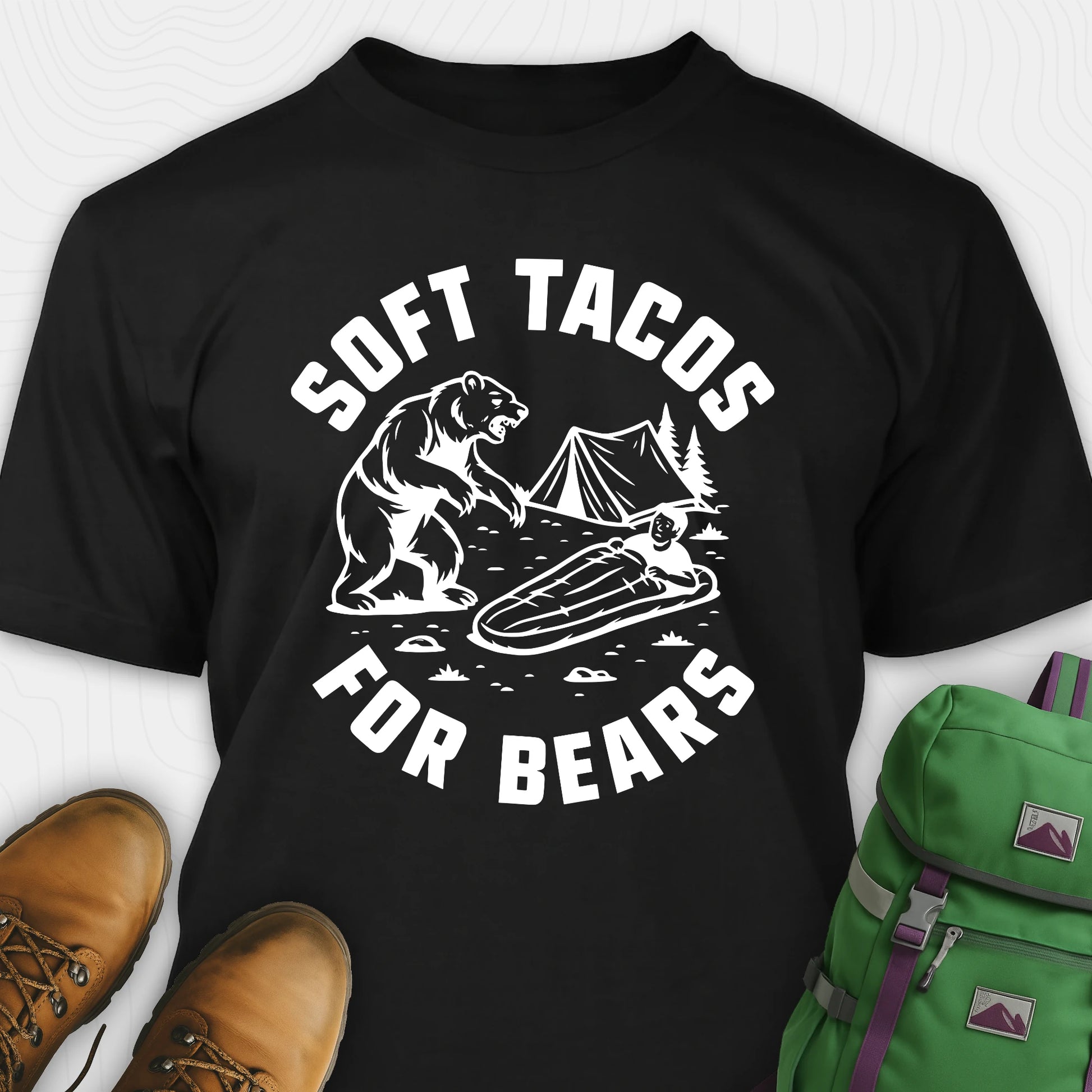 Black funny bear shirt with bear graphic and text 'Soft Tacos for Bears' on a white background. surrounded by hiking boots and a green backpack.