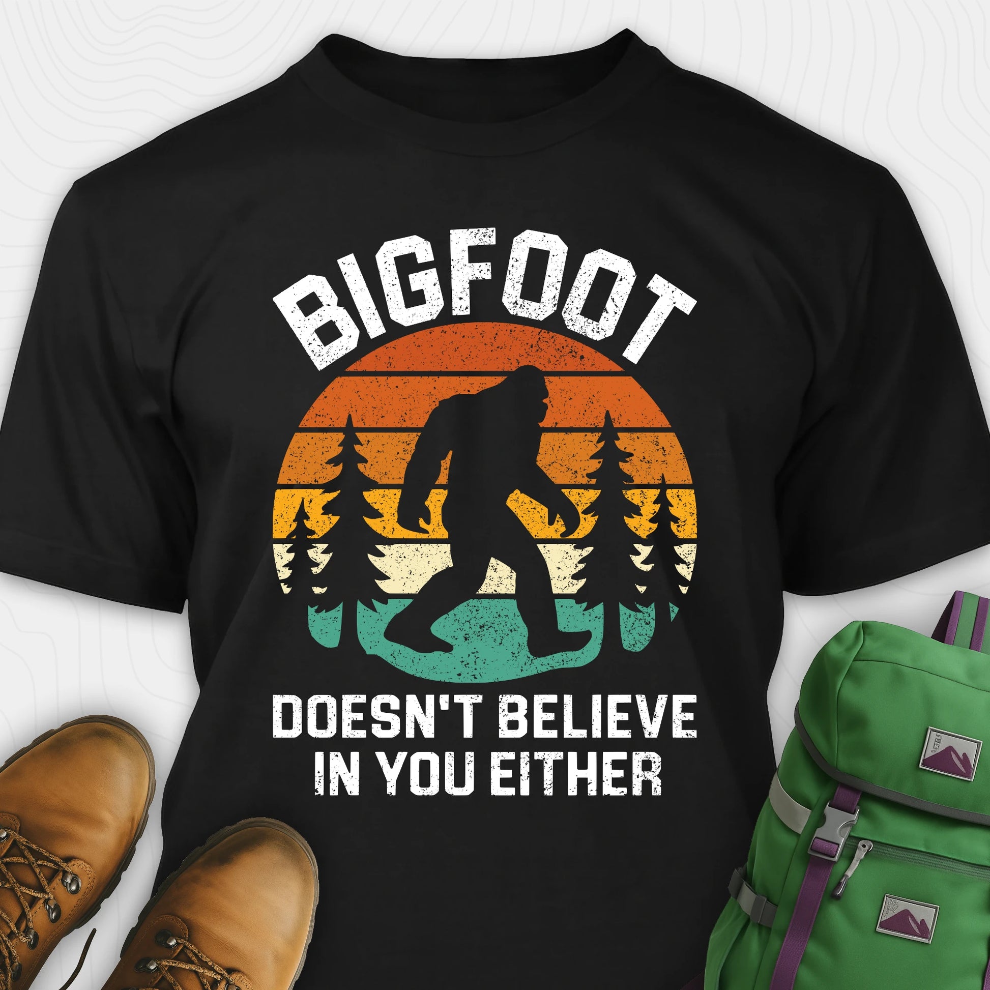Black funny Bigfoot shirt with sasquatch silhouette and humorous saying, paired with brown boots and a green backpack on a white background.