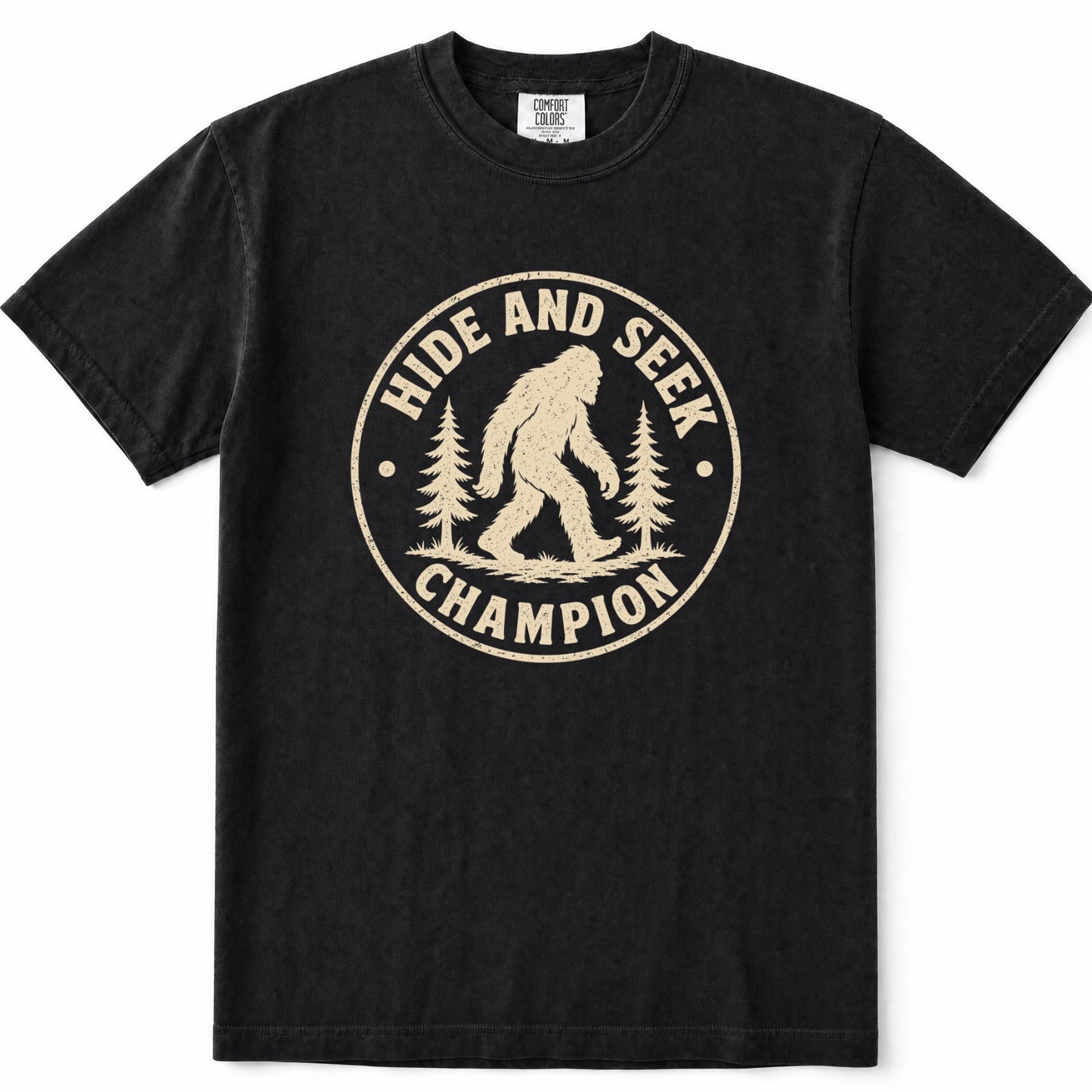 Black short-sleeve crewneck t-shirt featuring a distressed beige circular graphic on the chest with a Bigfoot silhouette and the words 'HIDE AND SEEK CHAMPION' alongside pine trees.