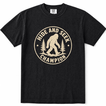 Black short-sleeve crewneck t-shirt featuring a distressed beige circular graphic on the chest with a Bigfoot silhouette and the words 'HIDE AND SEEK CHAMPION' alongside pine trees.