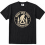Black short-sleeve crewneck t-shirt featuring a distressed beige circular graphic on the chest with a Bigfoot silhouette and the words 'HIDE AND SEEK CHAMPION' alongside pine trees.