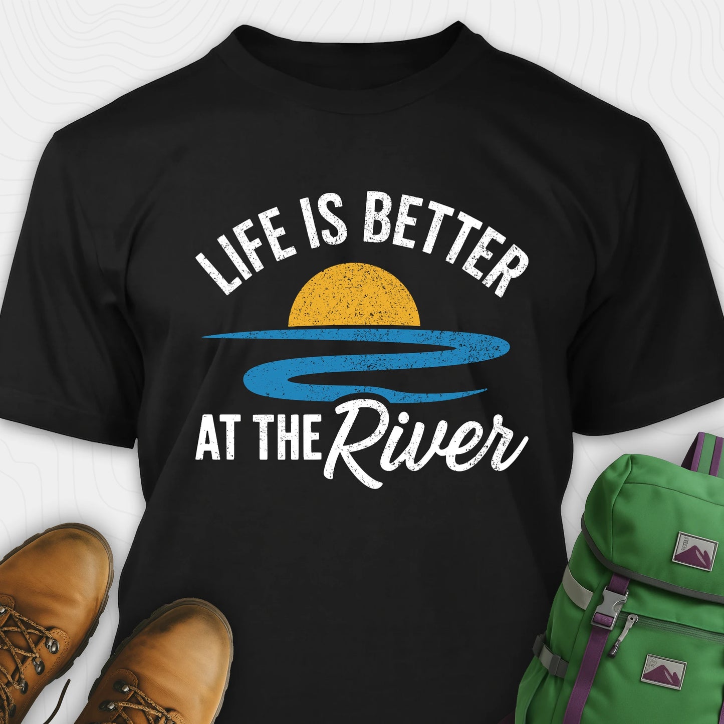 Black river shirt with 'Life is Better at the River' design, paired with brown boots and a green backpack on a light gray background.