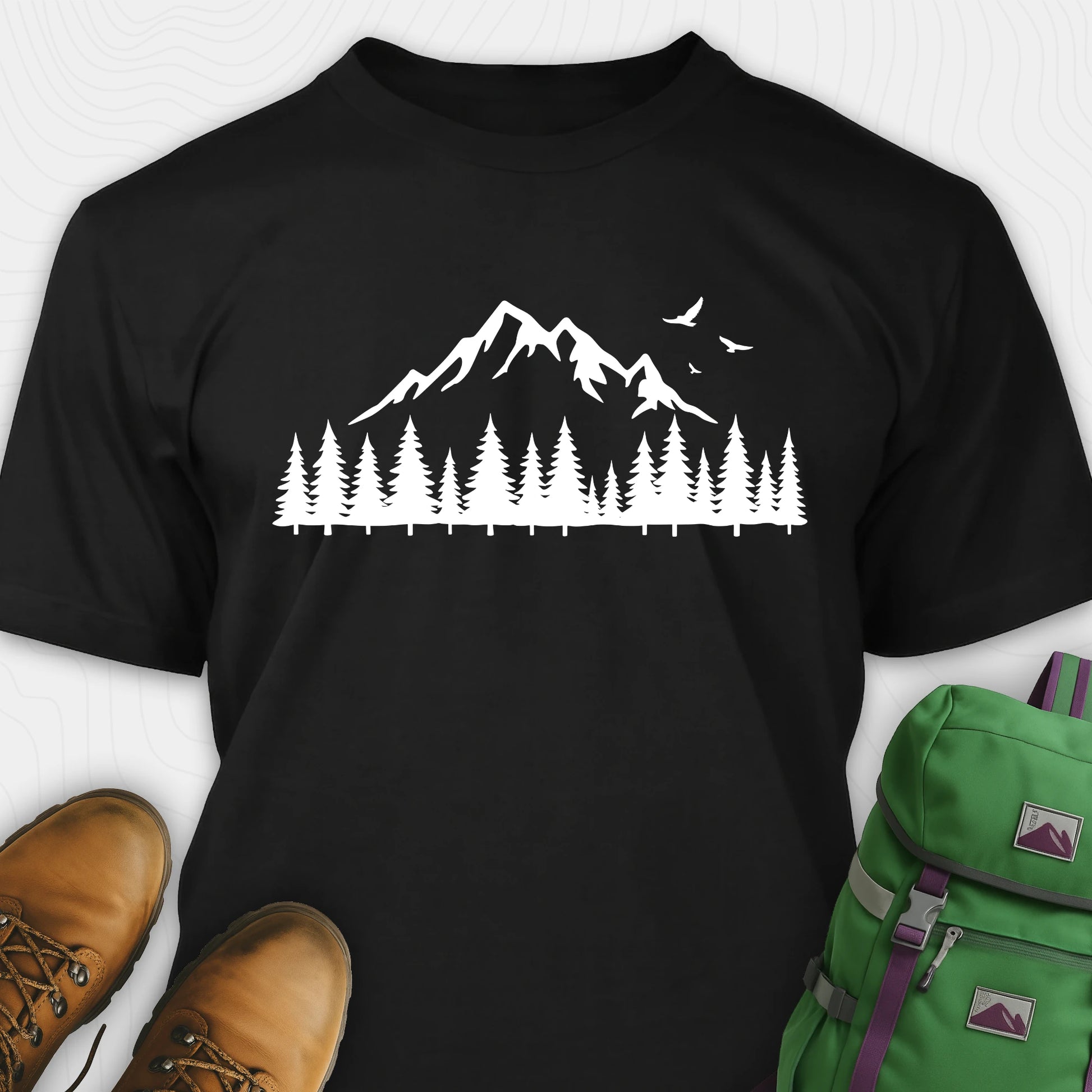 Black wild t-shirt with a minimalist mountain and pine tree graphic, shown flat with boots and a backpack