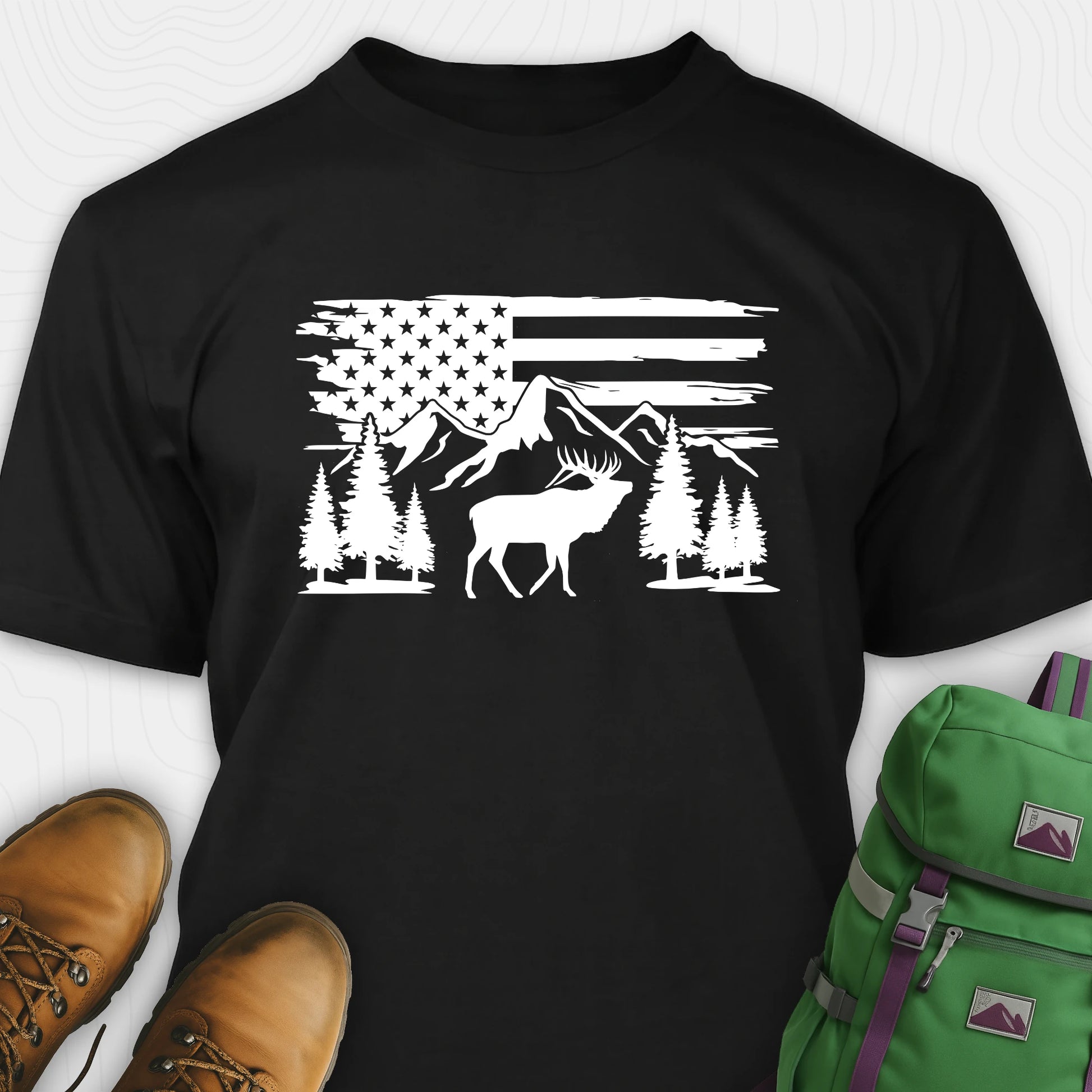 Black deer t-shirt with black graphic design of an American flag, trees, and a moose, accompanied by brown boots and a green backpack.