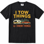 Black short-sleeve crewneck t-shirt with a vintage-style graphic print. The graphic features the text 'I TOW THINGS' in yellow, a vintage camper trailer and orange pickup truck towing pine trees, and '& I KNOW THINGS' in orange.