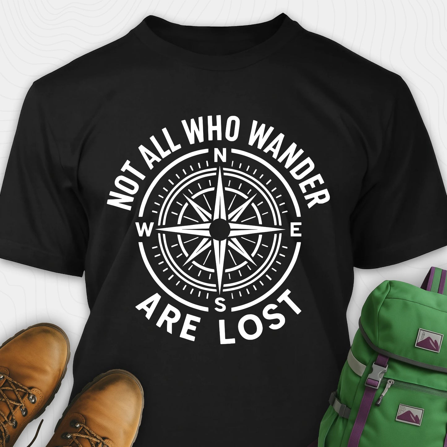 Black Not All Who Wander Are Lost T-shirt with compass design and text, paired with brown boots and a green backpack on a white background.