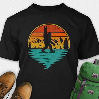 Black bigfoot shirt for men with retro mountain graphic beside boots and green backpack