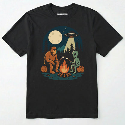 Black bigfoot and alien shirt, featuring a sasquatch and an alien roasting marshmallows around a campfire with moon and ufo graphic on a white background.