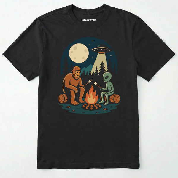 Black bigfoot and alien shirt, featuring a sasquatch and an alien roasting marshmallows around a campfire with moon and ufo graphic on a white background.