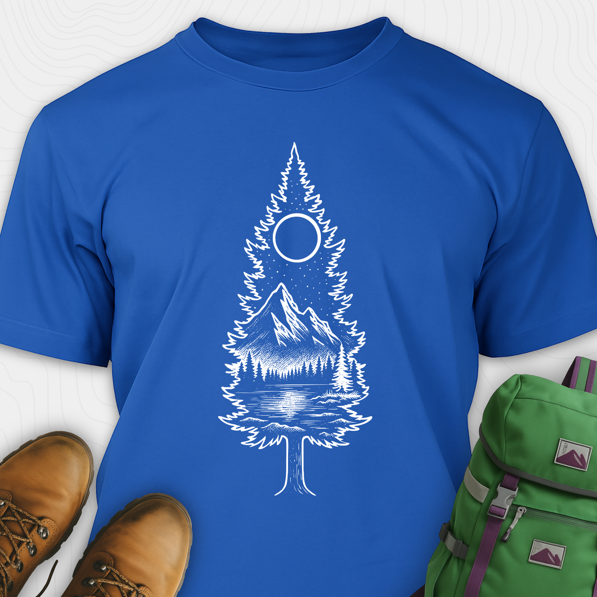 A blue tree t-shirt featuring a detailed white graphic of a pine tree silhouette. The tree outline contains a landscape scene: a mountain peak rising over a forested shoreline, with its reflection visible in a lake below. Above the mountain is a starry night sky and a full or partial circle representing the moon or sun. The t-shirt is displayed flat with brown leather boots to the lower left and a green and purple backpack to the lower right, suggesting an outdoor or hiking theme.