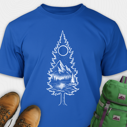 A blue tree t-shirt featuring a detailed white graphic of a pine tree silhouette. The tree outline contains a landscape scene: a mountain peak rising over a forested shoreline, with its reflection visible in a lake below. Above the mountain is a starry night sky and a full or partial circle representing the moon or sun. The t-shirt is displayed flat with brown leather boots to the lower left and a green and purple backpack to the lower right, suggesting an outdoor or hiking theme.