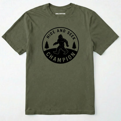 Military Green bigfoot hide and seek champion shirt with forest emblem on a white background.