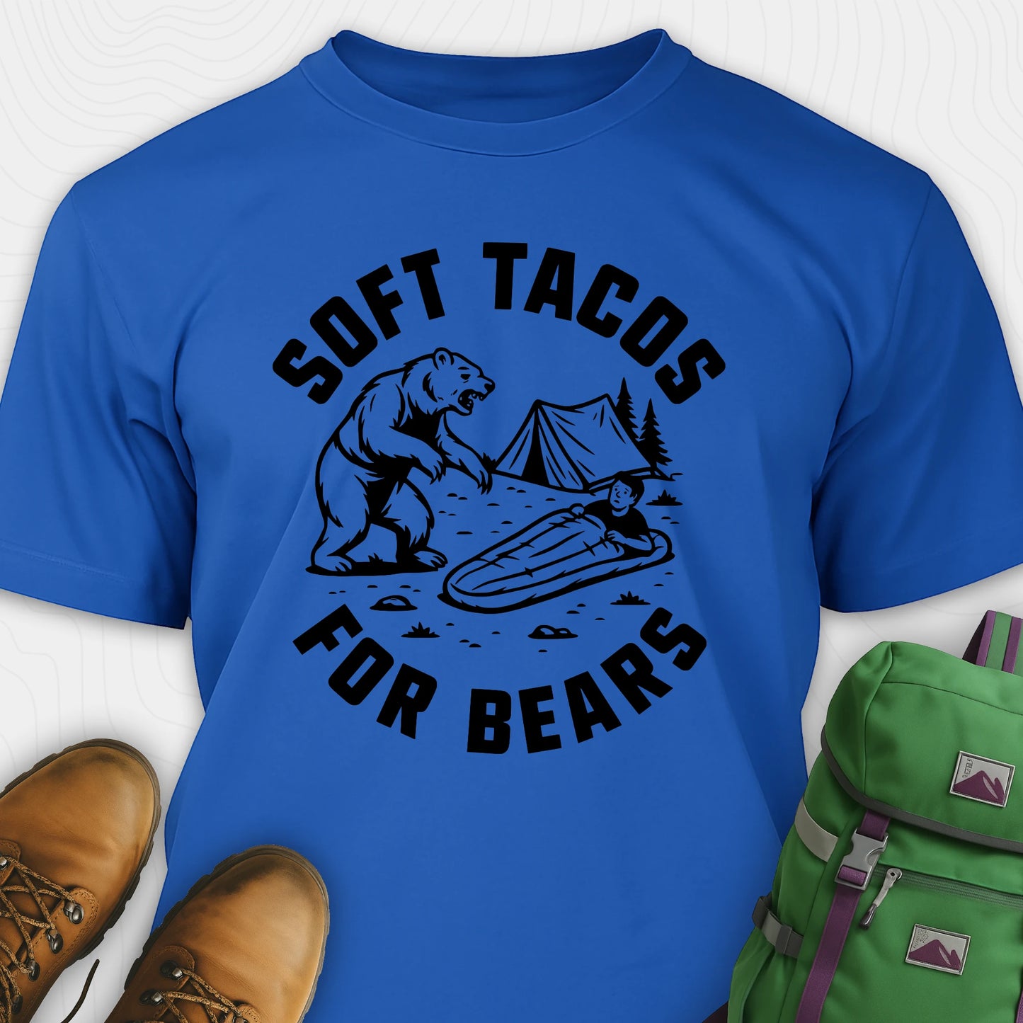 Blue funny bear shirt with bear graphic and text 'Soft Tacos for Bears' on a white background. surrounded by hiking boots and a green backpack.