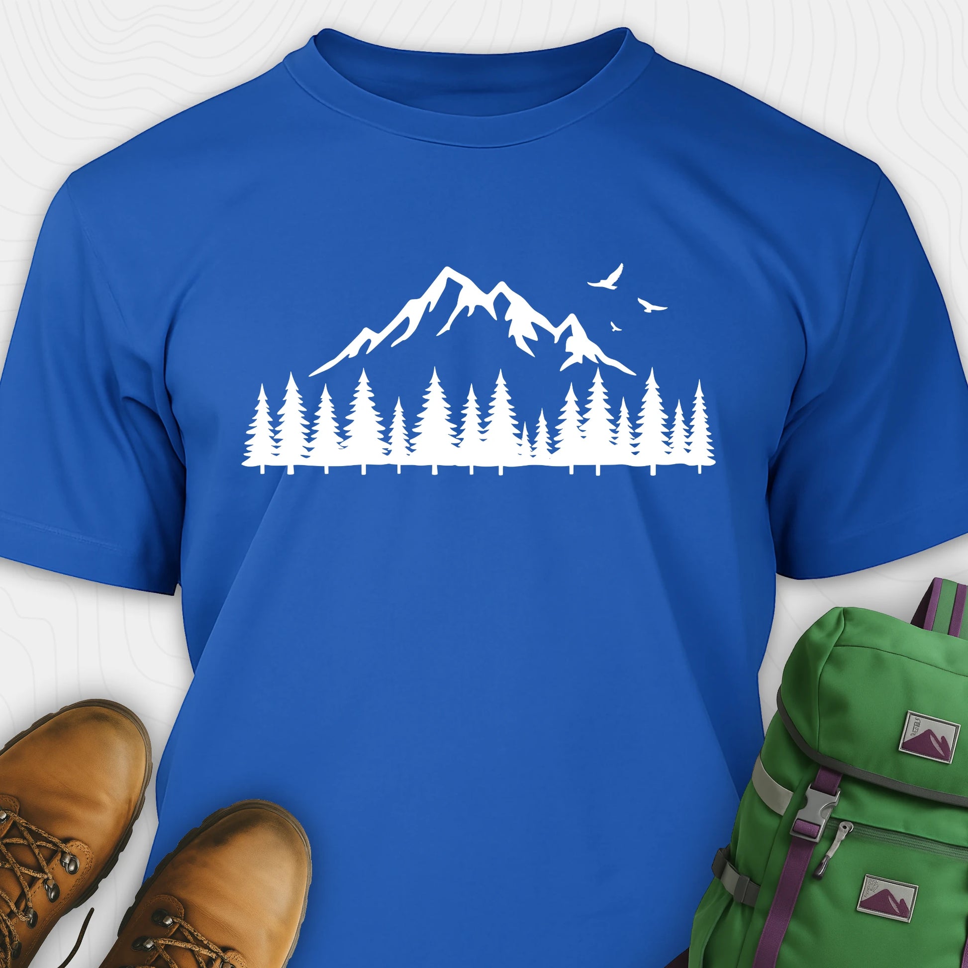 Blue wild t-shirt with a minimalist mountain and pine tree graphic, shown flat with boots and a backpack