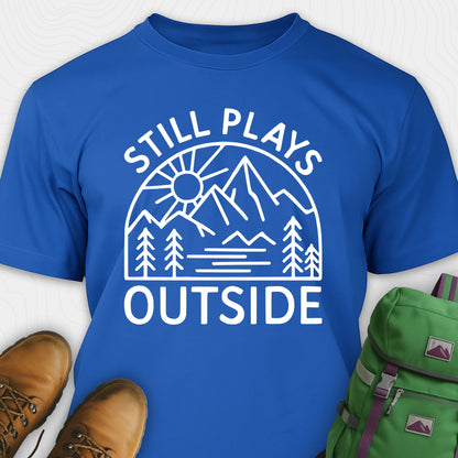Blue t-shirt with 'Still Plays Outside' graphic design, paired with brown boots and a green backpack on a light gray background.