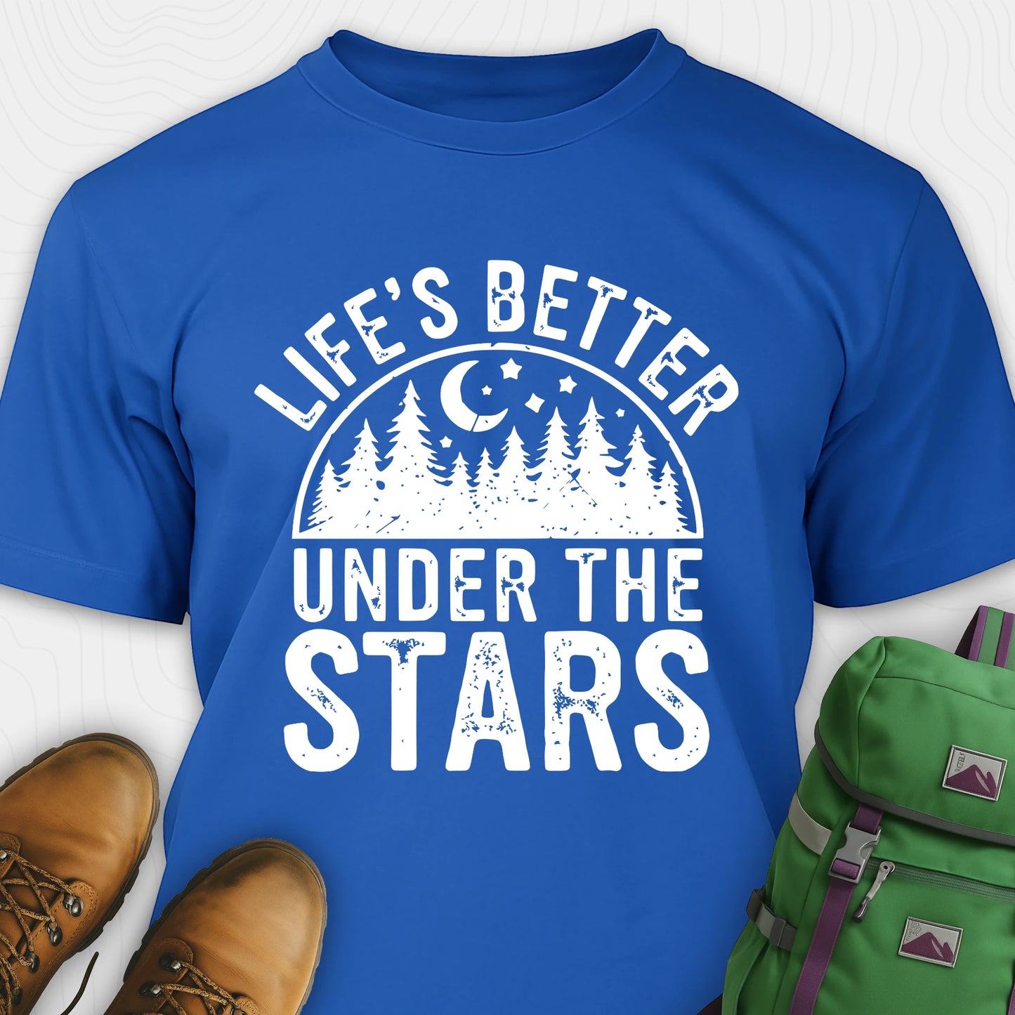 Royal blue Life's Better Under the Stars t-shirt with forest and moon graphic, paired with brown boots and a green backpack on a white background.