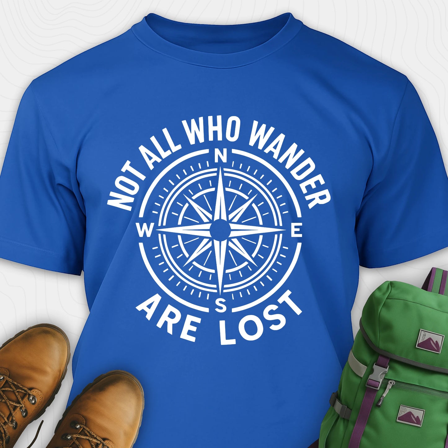 Blue Not All Who Wander Are Lost T-shirt with compass design and text, paired with brown boots and a green backpack on a white background