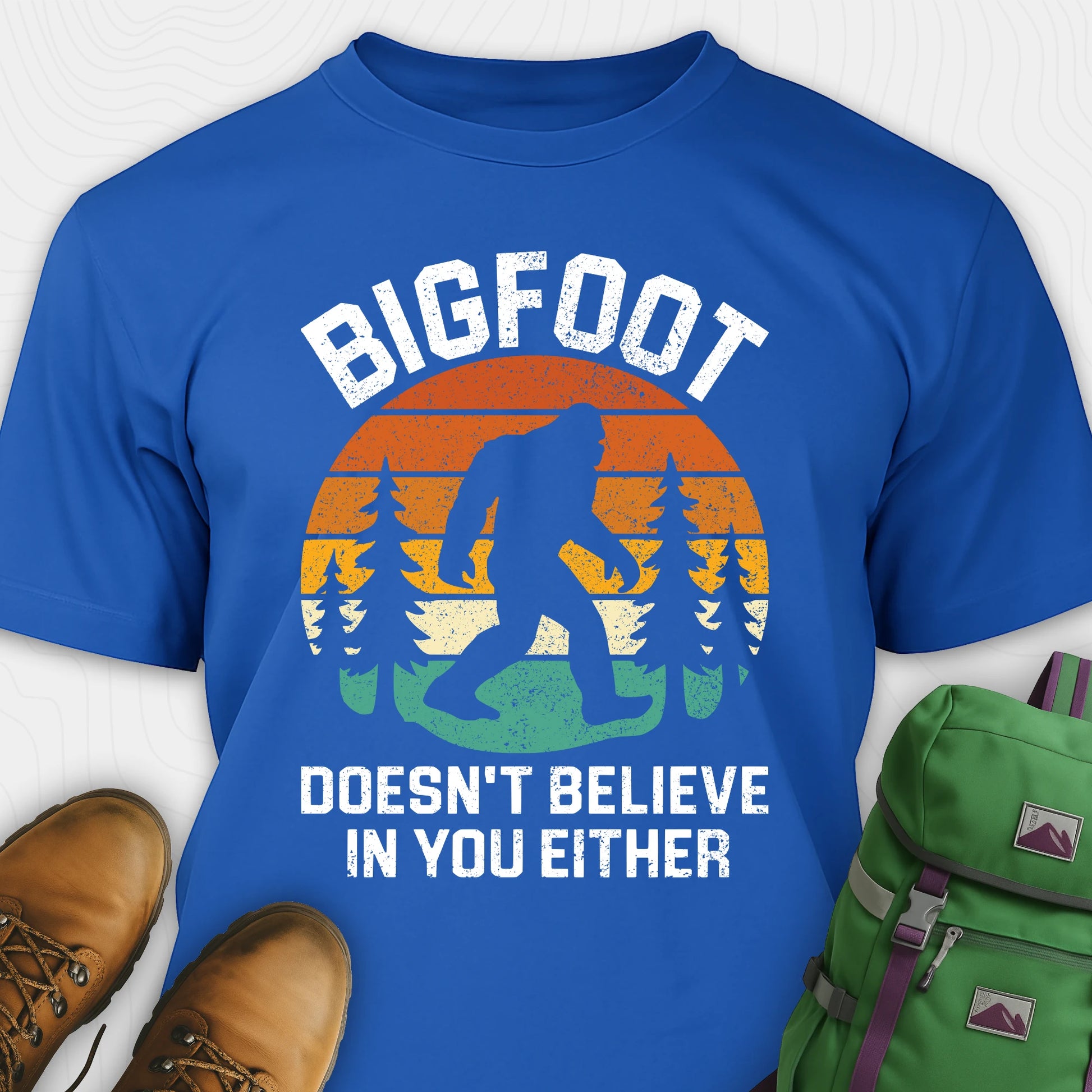 Blue funny Bigfoot shirt with sasquatch silhouette and humorous saying, paired with brown boots and a green backpack on a white background.