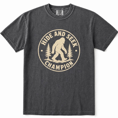 Pepper mineral-wash t-shirt with a distressed beige graphic on the front that reads 'HIDE AND SEEK CHAMPION' around an illustration of a Bigfoot figure and pine trees.